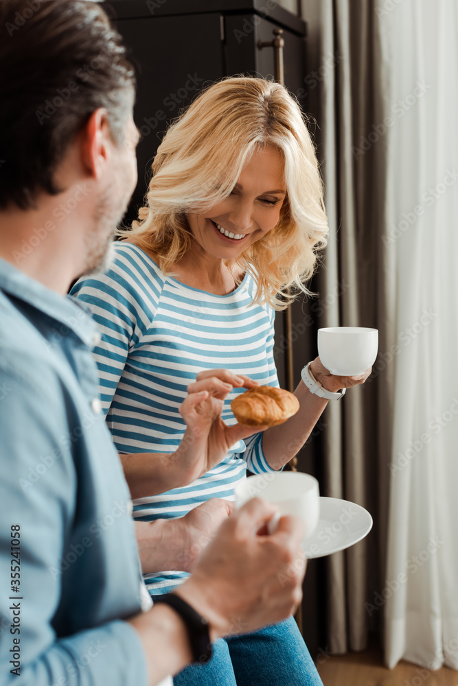 Selective focus of smiling woman holding croissant near husband with coffee in kitchen Selective focus of smiling woman holding croissant near husband with coffee in kitchen