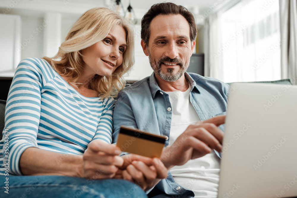 Selective focus of smiling man pointing at laptop near wife holding credit card in living room Selective focus of smiling man pointing at laptop near wife holding credit card in living room