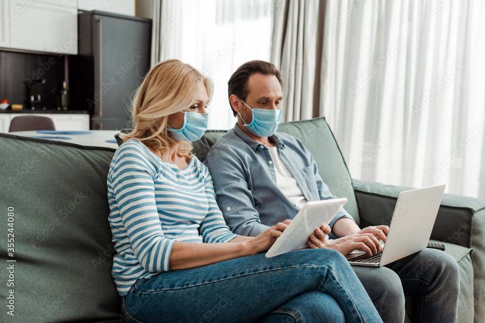 Mature couple in medical masks using laptop and digital tablet on couch Mature couple in medical masks using laptop and digital tablet on couch