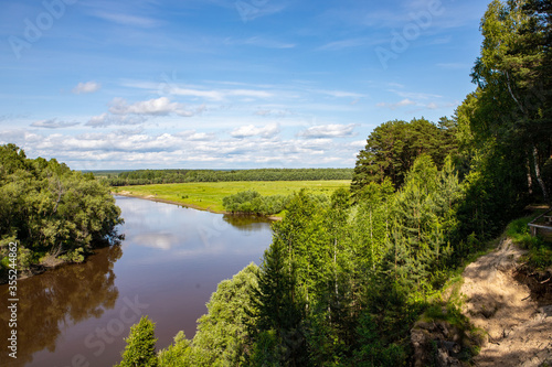 A small river flows around the forest, with the reflection of the trees in it against the blue sky. Nature of the Siberian expanses.