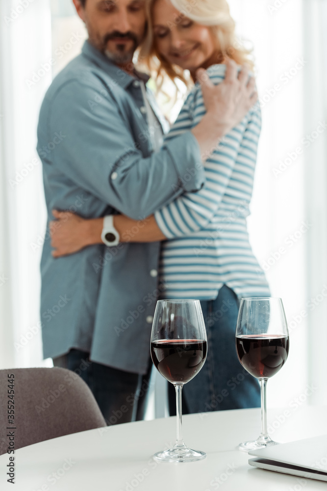Selective focus of glasses of wine and laptop on table near smiling mature couple embracing in kitchen