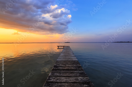 Old wooden jetty  pier reveals views of the beautiful lake  blue sky with cloud. Sunrise enlightens the horizon with orange warm colors. Summer landscape. Free space for text.