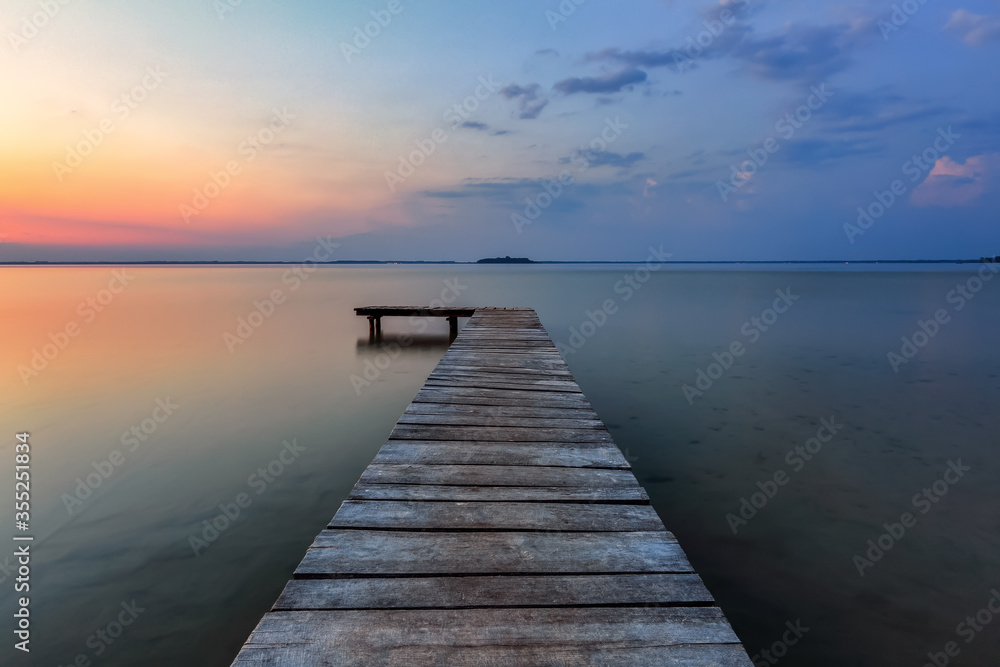 Old wooden jetty, pier reveals views of the beautiful lake, blue sky with cloud. Sunrise enlightens the horizon with orange warm colors. Summer landscape. Free space for text.