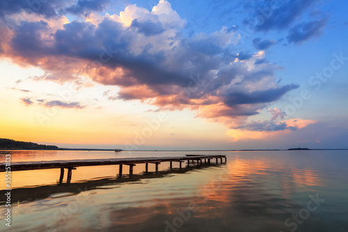 Fototapeta Naklejka Na Ścianę i Meble -  Old wooden jetty, pier reveals views of the beautiful lake, blue sky with cloud. Sunrise enlightens the horizon with orange warm colors. Summer landscape. Free space for text.