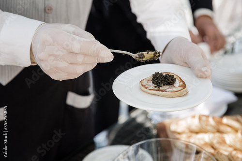 The waiter smears the pancakes with black caviar. A white plate in his hands. 