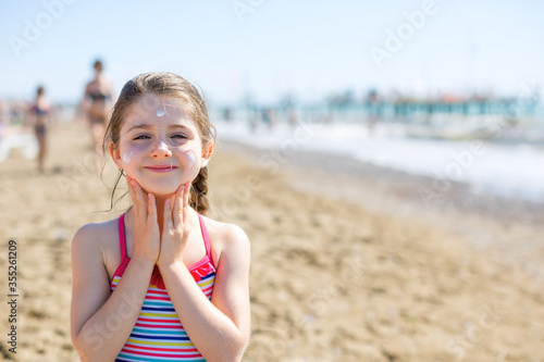 Cute little girl smiling and applying sunscreen on the face on the beach - summer vacation