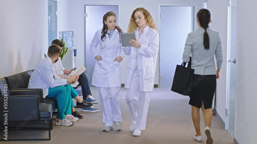 In the modern hospital corridor the main doctor female explain using a digital tablet to her colleague the diagnostic of the patient while walking in front of the camera In the modern hospital corridor the main doctor female explain using a digital tablet to her colleague the diagnostic of the patient while walking in front of the camera