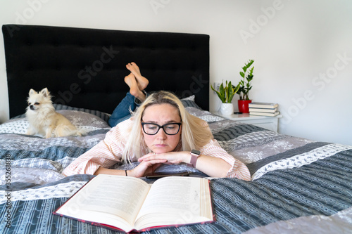 Foto Blonde hair woman sitting on cozy bed with dog, holding open book and reading