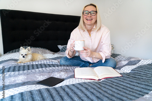 Foto Blonde hair woman sitting on cozy bed with dog, holding open book and reading
