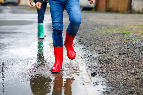 Close-up of legs of two kids boys wearing red and green rain boots and walking during sleet and rain on rainy cloudy day. Children jumping into puddle. Having fun outdoors, healthy children activity