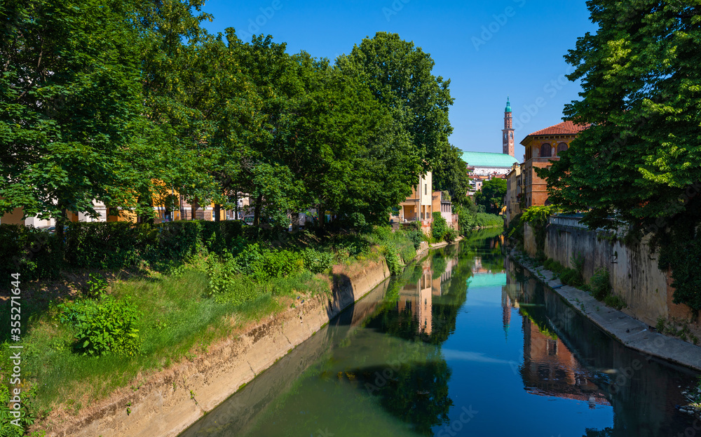 Fototapeta premium The stretch of the Retrone river that crosses the city of Vicenza, runs along Piazza dei Signori, the Palladian Basilica and passes under the Ponte San Michele. Big trees, a clear spring day, Italy.
