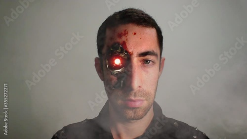 Cinemagraph of dramatic closeup portrait of a young bearded man with an expressionless face and revealed half cyborg humanoid face looking at the camera. He is with serious facial expressions. Loop.