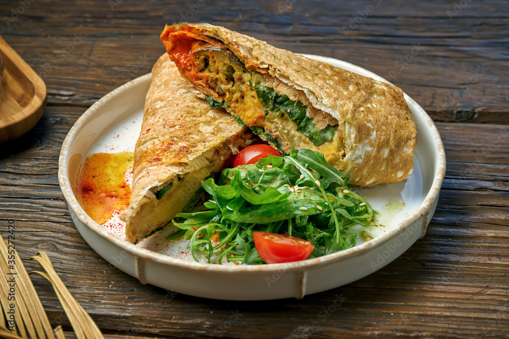 Vegetarian shawarma roll with spinach, tomatoes, hummus and melted cheese in a plate on a wooden background Vegetarian shawarma roll with spinach, tomatoes, hummus and melted cheese in a plate on a wooden background