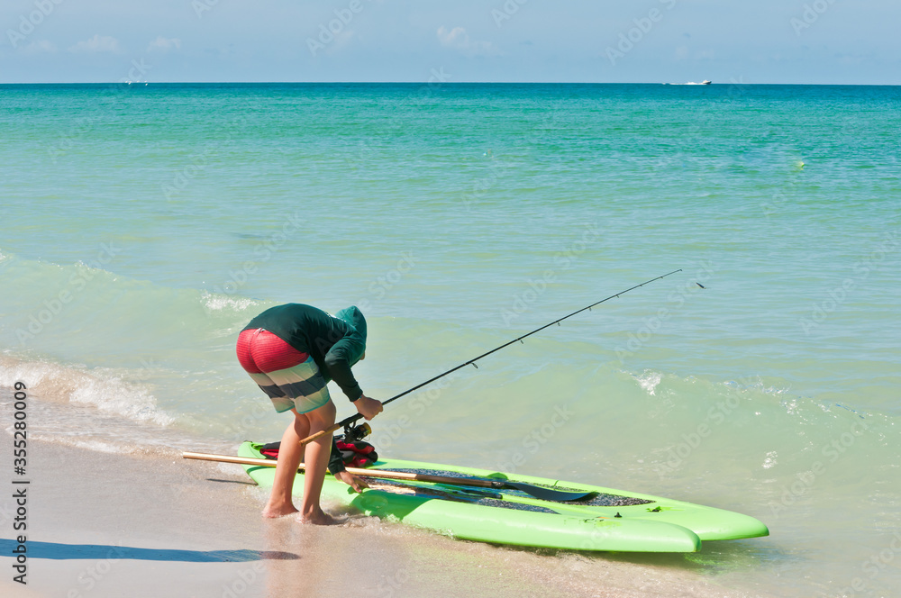Back view, medium distance of a male teenager with a fishing rod and green paddle board, preparing to go fishing off a sandy beach on gulf of Mexico on sunny day