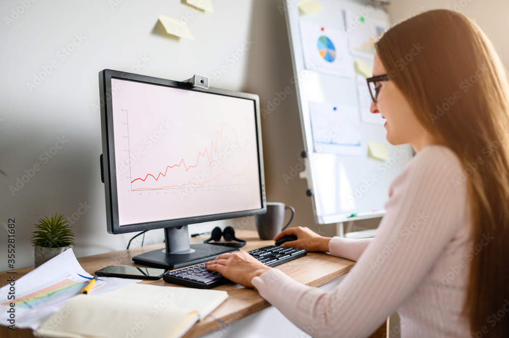 Young woman is using PC for work from home office, woman in eyeglasses looks at computer screen with a graphs on it Young woman is using PC for work from home office, woman in eyeglasses looks at computer screen with a graphs on it