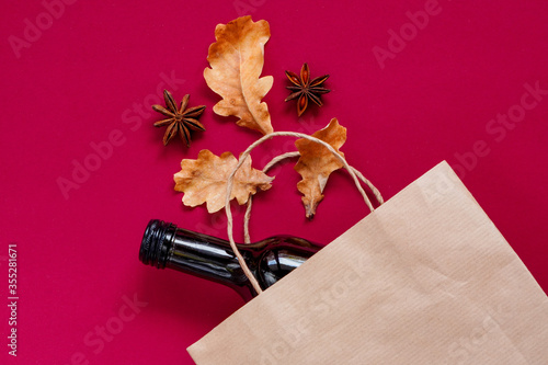 Paper bag with dark glass bottle of wine, alcohol present with dry leaves, composition. Hot vine aet for autumn. Flatly on maroon background, zero waste. Garbage recycling