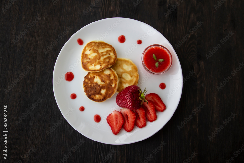Three pancakes with cheese and strawberries and sauce on a white dish against wood background. Delicious cheese pancake and strawberries for the breakfast or lunch. Tasty meal with berries. Three pancakes with cheese and strawberries and sauce on a white dish against wood background. Delicious cheese pancake and strawberries for the breakfast or lunch. Tasty meal with berries.