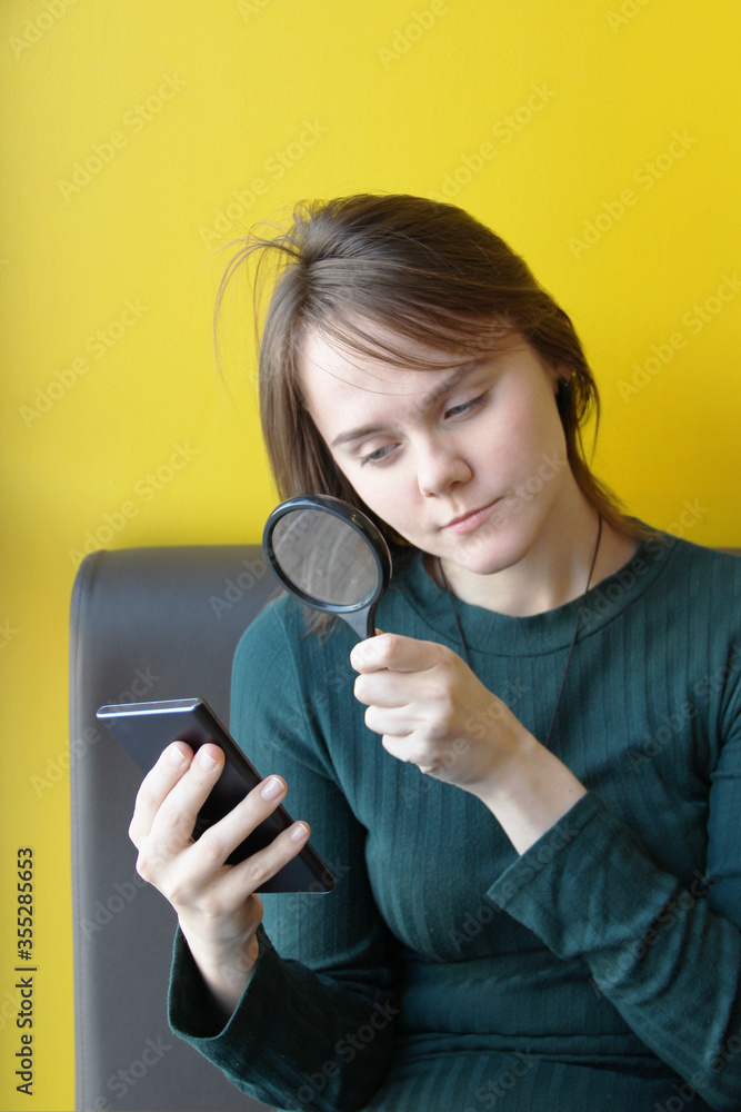 A young beautiful girl of European appearance in a blue jacket sits in a cafe with a mobile phone on the couch against the background of a yellow wall. Looks through a magnifying glass at the gadgets A young beautiful girl of European appearance in a blue jacket sits in a cafe with a mobile phone on the couch against the background of a yellow wall. Looks through a magnifying glass at the gadgets
