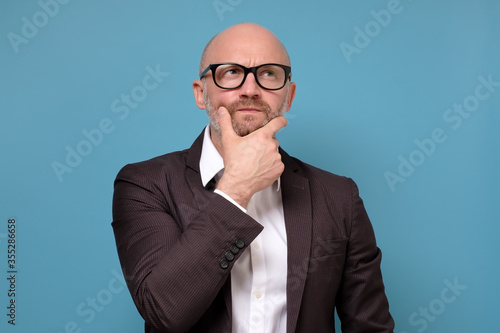 Thoughtful suspicious mature man looking up feeling skeptic doubtful. Businessman thinking holding hand on chin isolated on blue studio background photo