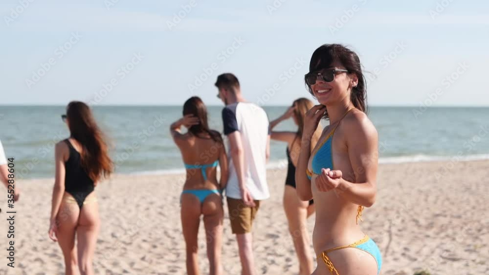 Pretty woman in bikini calls to swim in sea with her looking at camera. Back view of girl in swimsuit walking on beach, turns and invites to join, follow. Friends go to the ocean shore in slow motion. Pretty woman in bikini calls to swim in sea with her looking at camera. Back view of girl in swimsuit walking on beach, turns and invites to join, follow. Friends go to the ocean shore in slow motion.