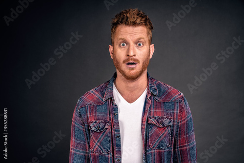 Redhead male with bristle dropping jaw as seeing incredible and amazing scenery, posing over gray background in casual shrit, gasping, staring thrilled and astonished at camera over grey wall
