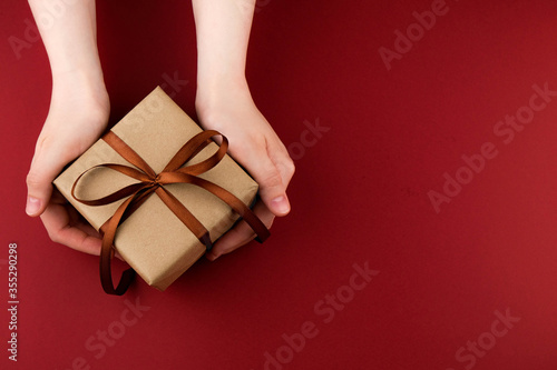 Female hands hold a gift box wrapped in craft paper and tied with a brown ribbon on a red background close-up. Copy space