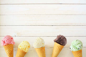 Ice cream cone bottom border with a variety of flavors. Top down view over a white wood background with copy space.