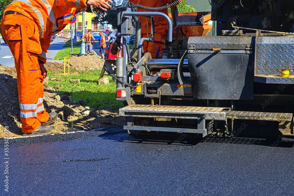Worker makes blacktop. New asphalt is laid over the old asphalt at the intersection, filling in several potholes in the road. Workers made repairs to the roadway at the intersection.