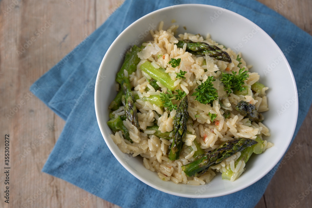 Risotto with green asparagus and parsley garnish in a white bowl, blue napkin on rustic wood with copy space, high angle view from above, selected focus