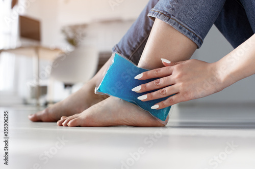 Close up woman feet and legs, and hands holding ice gel pack as cold compress on ankle due to stretching or injury, sitting on floor in apartment indoors photo