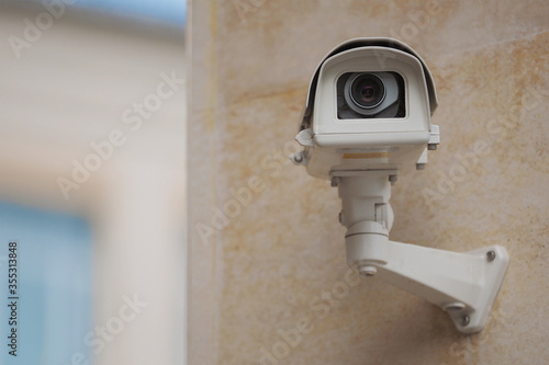 Big white surveillance camera on light beige stone column against background of public building with blue window; close-up