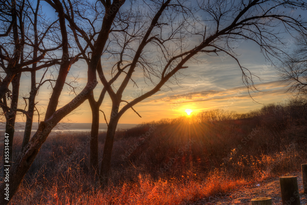 sunset sending rays across a leafless, winter forest with the Mississippi River in the distance, view from bluff, tree silhouettes framing a colorful sky, Pierre Marquet State Park Illinois sunset sending rays across a leafless, winter forest with the Mississippi River in the distance, view from bluff, tree silhouettes framing a colorful sky, Pierre Marquet State Park Illinois