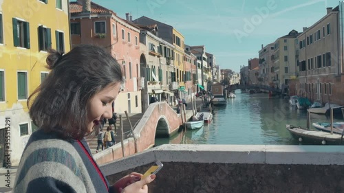 Beautiful asian woman tourist in red sweater enter in frame taking photo of the picturesque venetian canal with her smartphone from edge of the bridge. Charming woman spending her vacation in Venice.