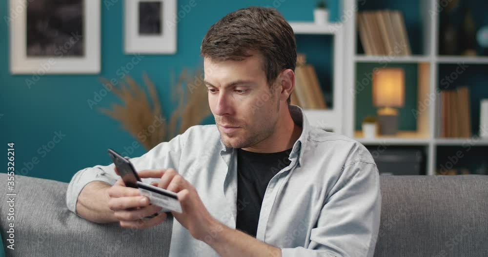 Handsome man in domestic outfit entering data from his credit card on smartphone for online payments. Concept of conveniences and modern technologies Handsome man in domestic outfit entering data from his credit card on smartphone for online payments. Concept of conveniences and modern technologies