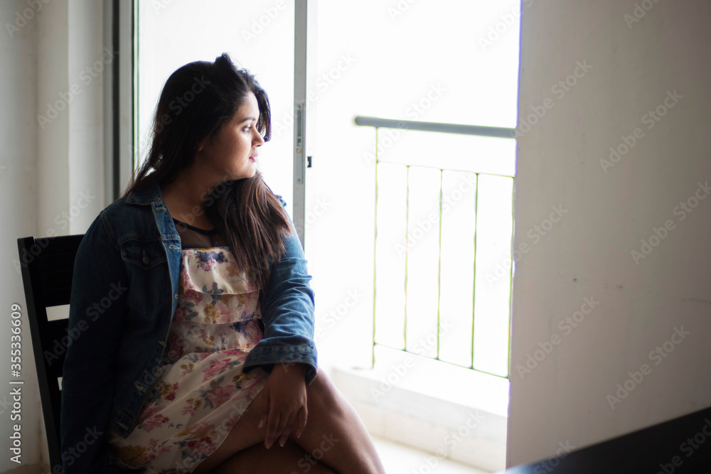 Indian Bengali beautiful and cute brunette girl in a casual blue jeans shirt and white top sitting on a chair inside a room facing a glass door with sidelight. Indian lifestyle and fashion portrait Indian Bengali beautiful and cute brunette girl in a casual blue jeans shirt and white top sitting on a chair inside a room facing a glass door with sidelight. Indian lifestyle and fashion portrait
