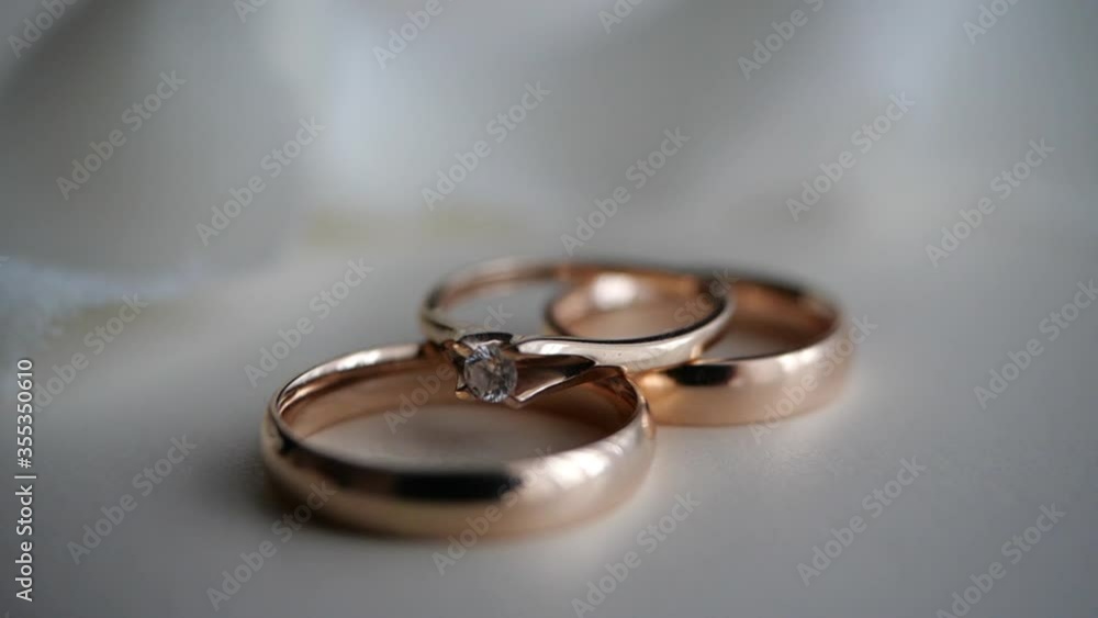 Three beautiful gold rings-a diamond engagement ring and an engagement ring-lie on a white table against a light fabric background. Gifts and romance, decor. Festive atmosphere of the bride and groom. Three beautiful gold rings-a diamond engagement ring and an engagement ring-lie on a white table against a light fabric background. Gifts and romance, decor. Festive atmosphere of the bride and groom.
