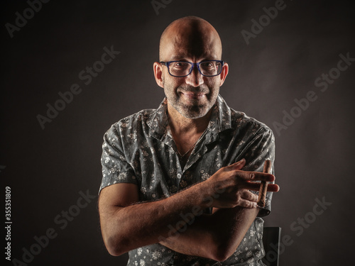 Portrait of a bald man with glasses, with a bristle man with a cigar, who is sitting on a chair in a dark space. photo