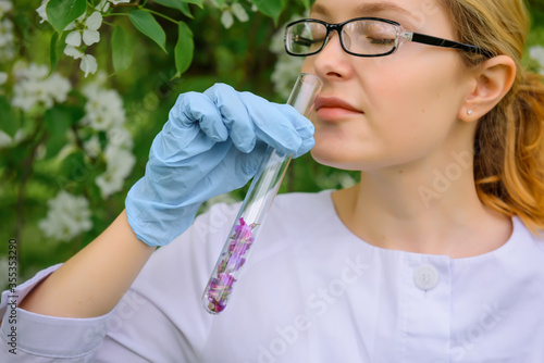 Beautiful female scientist in medical gloves inhales aroma from test tube with flower petals on the background of blooming tree in botanical garden  close-up. Creation of a perfume  natural cosmetics.