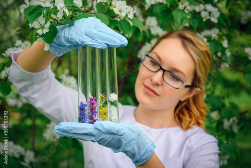 Beautiful female scientist in medical gloves holds test tubes with flower petals on the background of blooming tree in botanical garden, close-up. Creation of a perfume fragrances, natural cosmetics.