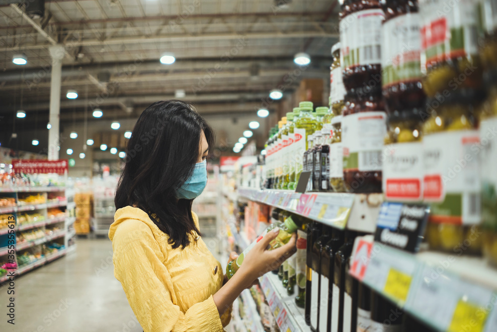 Asian woman wearing face mask buying in supermarket. Young female panic shopping on shelf during Coronavirus covid-19 pandemic. Women shopping reading product label.