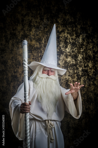 A grey-haired bearded severe wizard in a white cloak and a cap with a staff against a golden textured background.