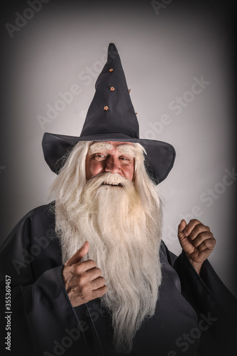 A stern grey-haired bearded wizard in a gray cassock and a cap is practicing sorcery and doing magic against a light background.