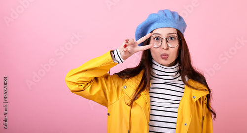 A young girl in a stylish beret hat and a yellow raincoat with glasses on a pink background.