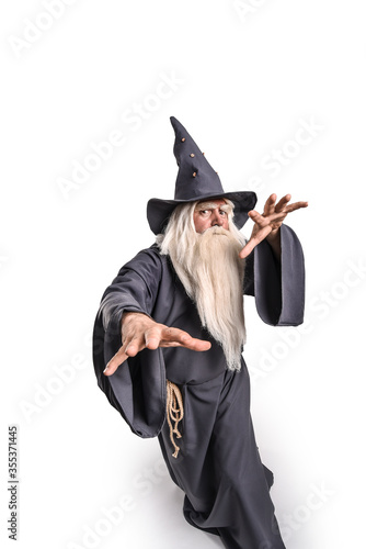 A stern grey-haired bearded wizard in a gray cassock and a cap is practicing sorcery and doing magic against a light background.