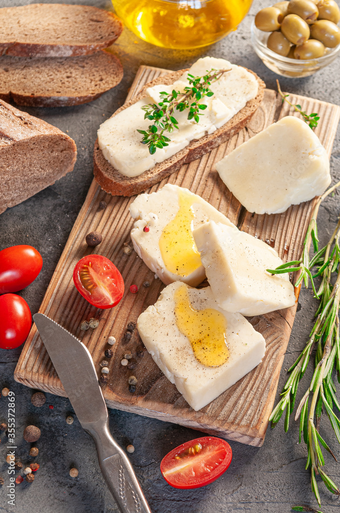 Adyghe cheese is cut into pieces. Cheese on a wooden board. Served with thyme, rosemary, spices and cherry tomatoes and olive oil. In the background are olives, rye bread and olive oil. Adyghe cheese is cut into pieces. Cheese on a wooden board. Served with thyme, rosemary, spices and cherry tomatoes and olive oil. In the background are olives, rye bread and olive oil.