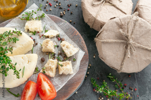 Adyghe cheese cut off a triangular piece. Cheese on a wooden board. Garnished with thyme, parsley, rosemary, tomatoes and spices. Next to the cheese in the package and olive oil.  photo