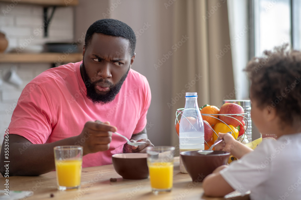 African american man in a pink tshirt feeding his kid and frowing to entertain her African american man in a pink tshirt feeding his kid and frowing to entertain her