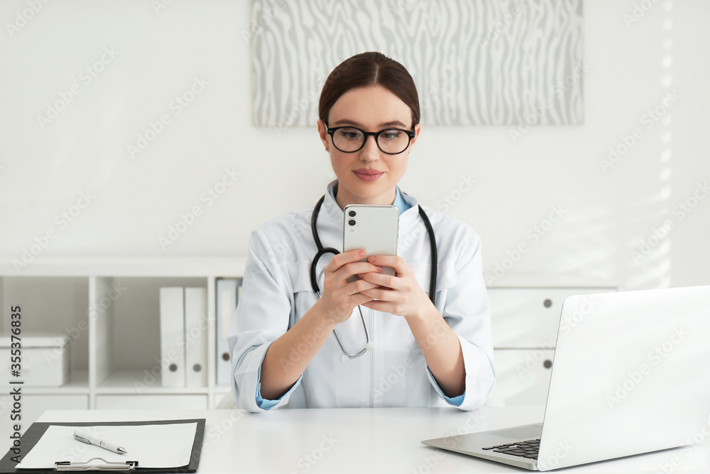 Young female doctor with smartphone at table in office Young female doctor with smartphone at table in office