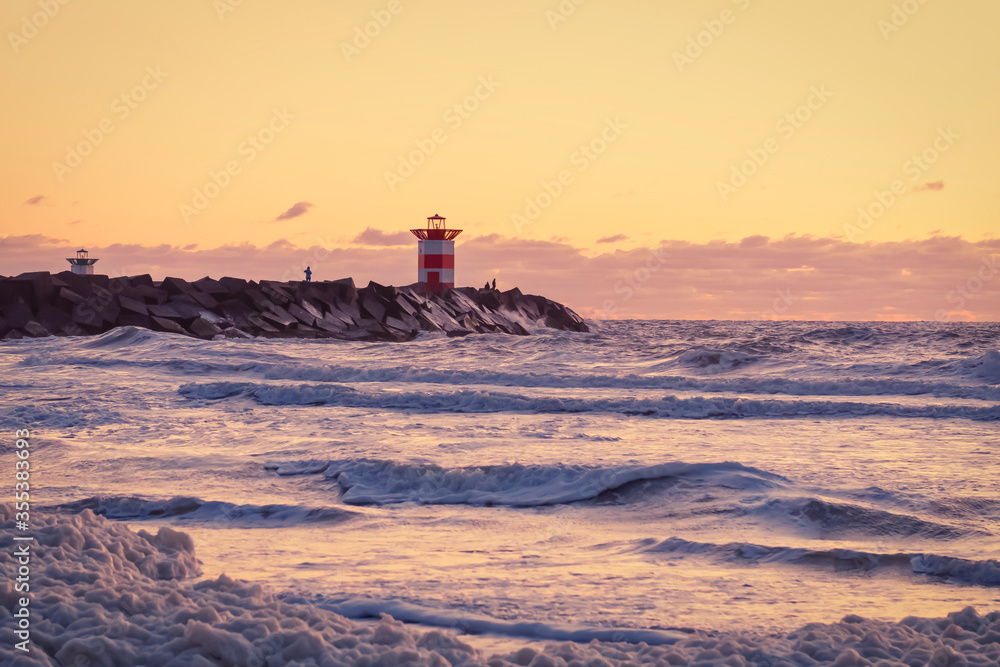 Red and white striped lighthouse during a vibrant sunset. Water hits the black boulders in waves. Dramatic sky in golden color. Scheveningen, the Netherlands