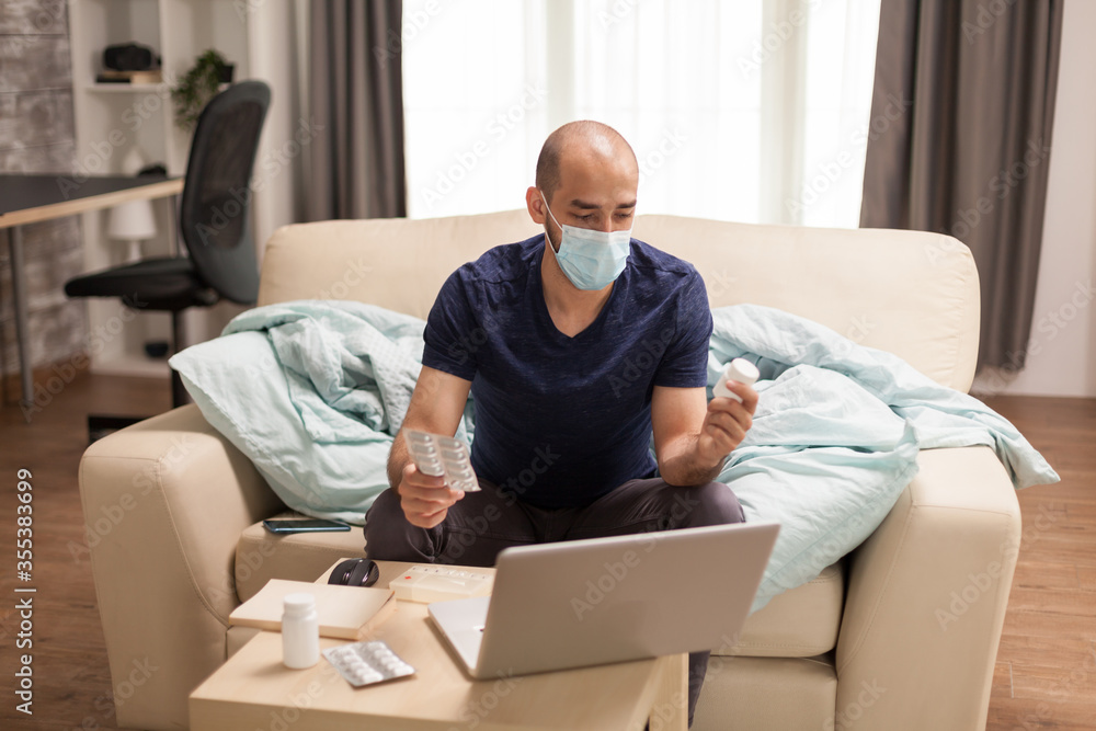 Sick man looking at pills bottle wearing protection mask during a video conference in time of self isolation. Sick man looking at pills bottle wearing protection mask during a video conference in time of self isolation.